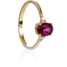 A product photo of a yellow gold grape garnet ring sitting against a white background. The gold band is plain and smooth, and the centre oval-cut warm purple garnet stone is framed by a single white diamond on either side.