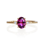 A product photo of a yellow gold grape garnet ring sitting against a white background. The gold band is plain and smooth, and the centre oval-cut warm purple garnet stone is framed by a single white diamond on either side.