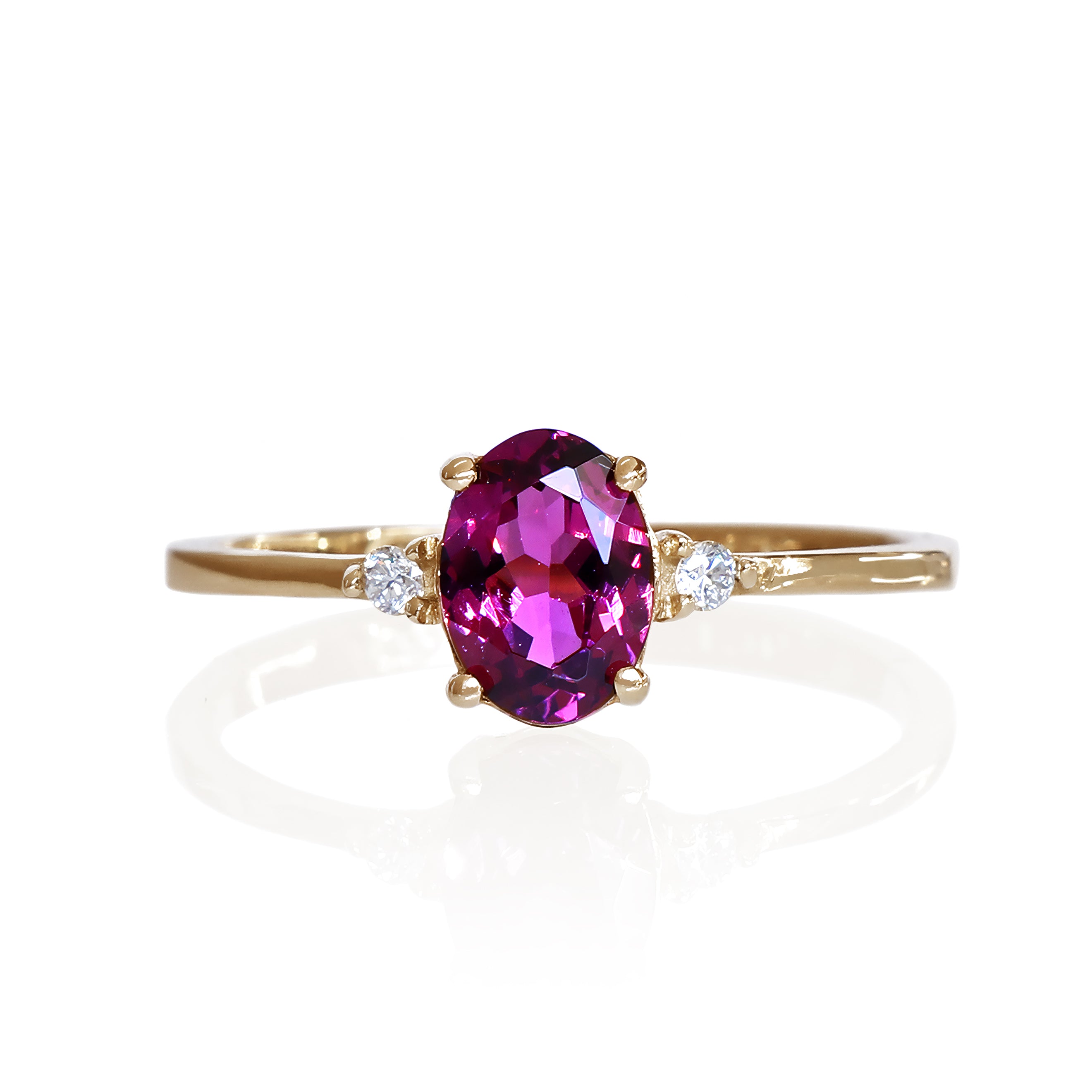 A product photo of a yellow gold grape garnet ring sitting against a white background. The gold band is plain and smooth, and the centre oval-cut warm purple garnet stone is framed by a single white diamond on either side.