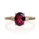 A product photo of a dainty and elegant yellow gold ring with a stunning trio of rhodalite and diamonds sitting against a white background. The largest of the stones sits in the centre, an oval rhodalite - hugged by two smaller circular white diamonds on either side.