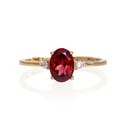 A product photo of a yellow gold red rhodalite ring sitting against a white background. The gold band is plain and smooth, and the centre oval-cut warm reddy pink rhodalite stone is framed by a single white diamond on either side.
