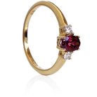 A product photo of a yellow gold rhodalite and diamond trio ring sitting on a white background. The 6x4mm oval purple rhodalite gemstone stands in stark contrast to the 0.03ct white diamond stones on either side.