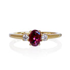 A product photo of a yellow gold rhodalite and diamond trio ring sitting on a white background. The 6x4mm oval purple rhodalite gemstone stands in stark contrast to the 0.03ct white diamond stones on either side.