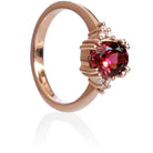 A product photo of a rose gold rhodalite and diamond trio ring sitting on a white background. The oval purple rhodalite gemstone stands in stark contrast to the little clusters of three white diamond stones on either side.