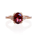 A product photo of a rose gold rhodalite and diamond trio ring sitting on a white background. The oval purple rhodalite gemstone stands in stark contrast to the little clusters of three white diamond stones on either side.