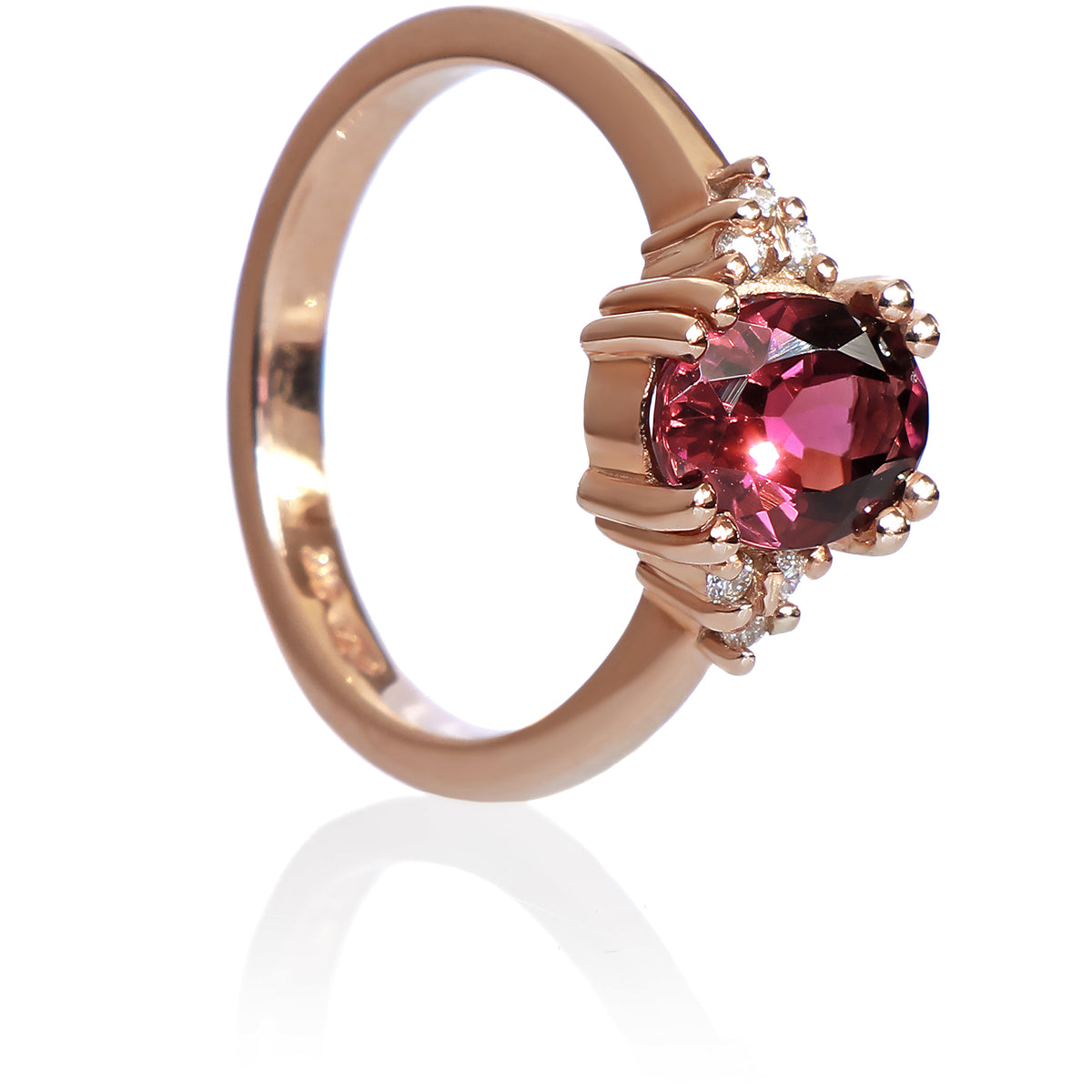 A product photo of a rose gold pink tourmaline and diamond trio ring sitting on a white background. The oval magenta pink tourmaline gemstone stands in stark contrast to the little clusters of three white diamond stones on either side.