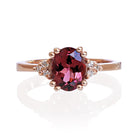 A product photo of a rose gold pink tourmaline and diamond trio ring sitting on a white background. The oval magenta pink tourmaline gemstone stands in stark contrast to the little clusters of three white diamond stones on either side.