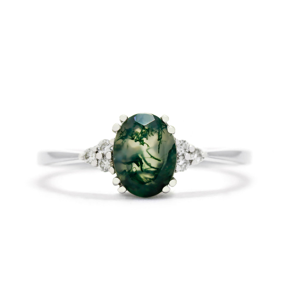 A product photo of a white gold moss agate and diamond trio ring sitting on a white background. The oval, naturally-included moss agate gemstone stands in stark contrast to the little clusters of three classic white diamond stones on either side.