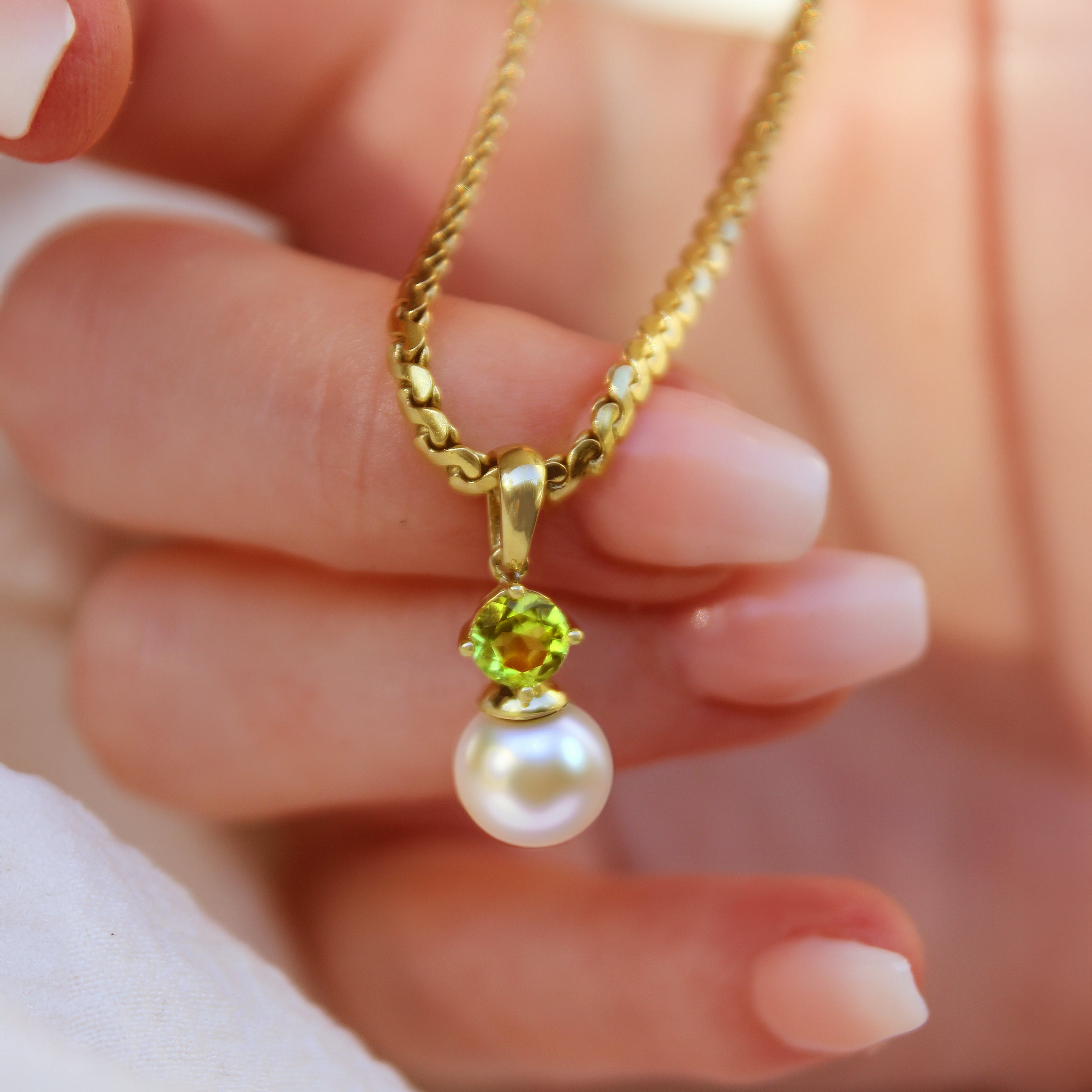 This product image is a close-up of a beautiful solid 9 karat yellow gold pearl and gemstone pendant design suspended by a gold chain. The pendant primarily features a round-cut bright green peridot stone in the centre, set with delicate gold claws above a single white round-shaped pearl. A thin gold chain connects to the pendant.