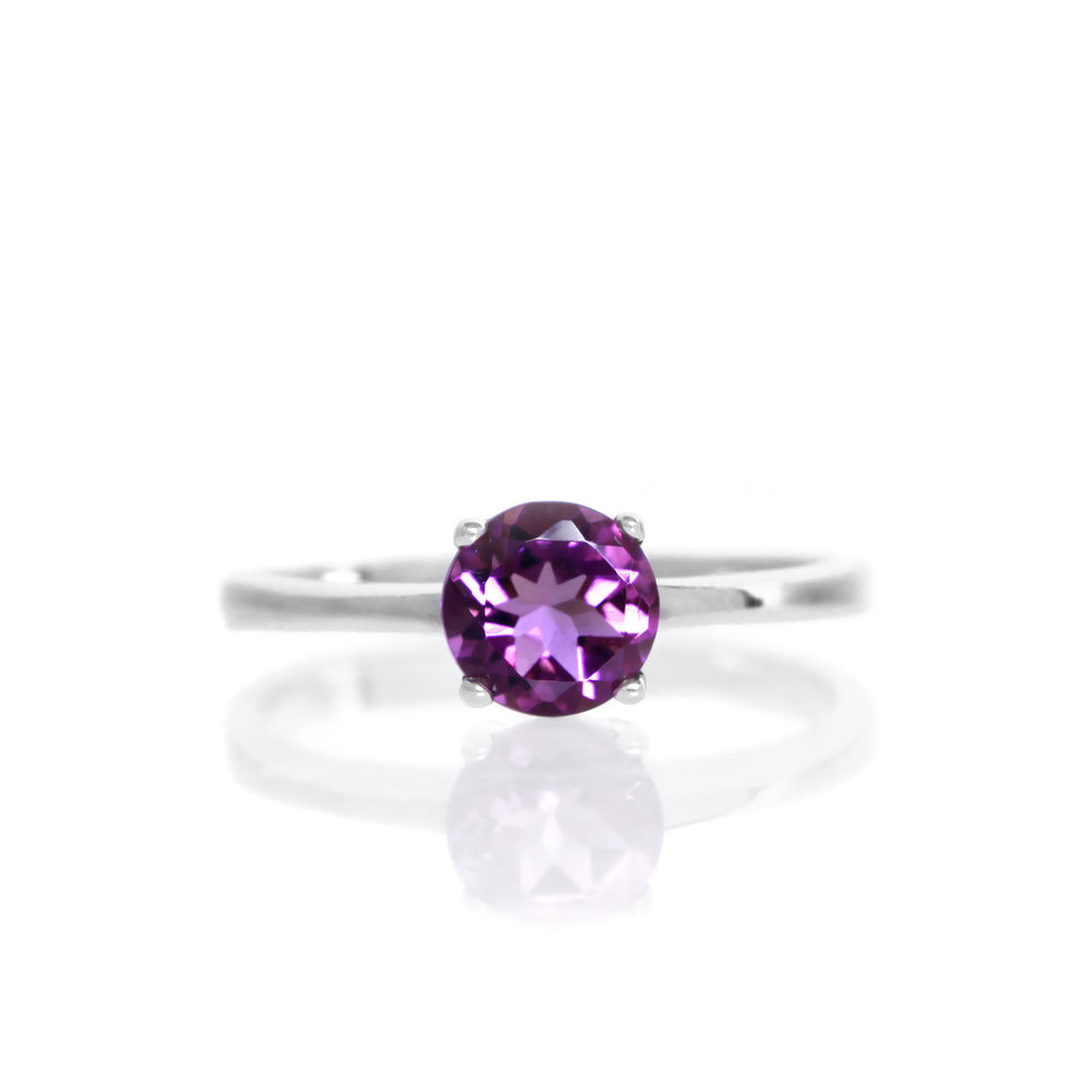 A product photo of a Round Amethyst Solitaire Ring in silver sitting on a plain white background. The amethyst centrestone measures 5.5mm across. The stone is a deep purple, and reflects violet and pink-hued light across its many faceted edges.
