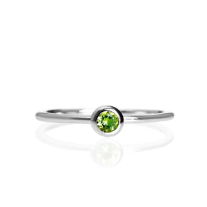 A product photo of a delicate white gold stacking ring with a tiny, bezel-set peridot in the centre sitting on a white background. The band is slim and thread-like, with the focus drawn to the petite 3mm glinting green centre stone.