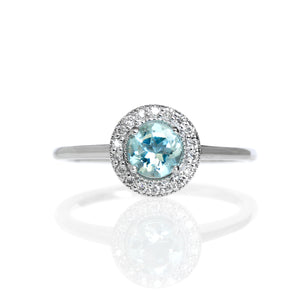 A product photo of a classic diamond halo aquamarine ring in solid 9k white gold on a white background. The diamond halo is framed by a thick band of solid 9k white gold, and the rounded band is smooth and minimalistic. The pale baby blue aquamarine centre stone is held in place by 4 claws and reflects light off of its many facets.