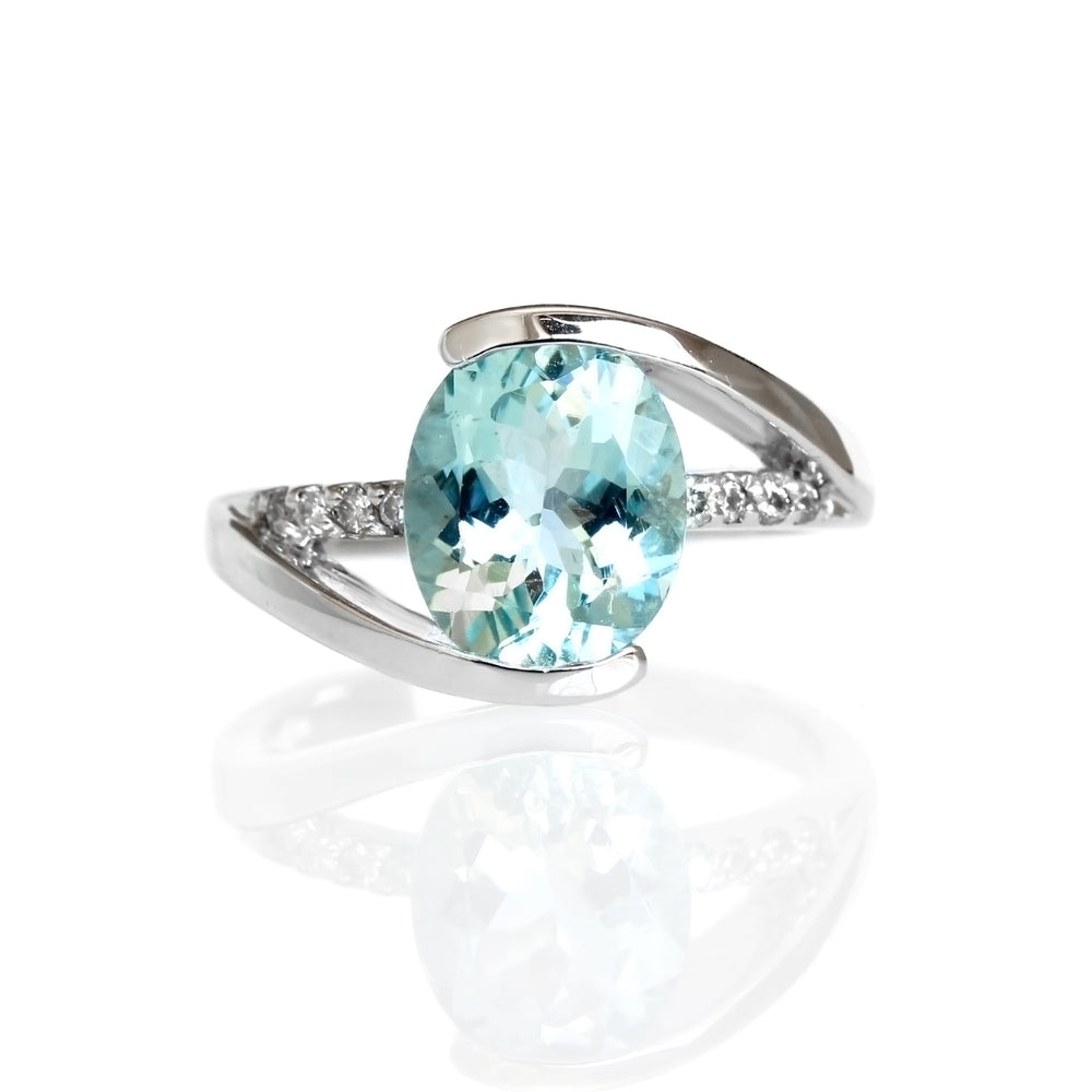 A product photo of a bold aquamarine and diamond statement ring in solid 9k white gold on a white background. The pale baby blue 10x8mm aquamarine centre stone is held in place by 4 claws and reflects light off of its many facets. The golden band splits in 3, two swooping to meet the top and bottom of the stone, and the third - decorated in diamond details - running straight through the centre.