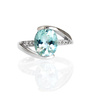 A product photo of a bold aquamarine and diamond statement ring in solid 9k white gold on a white background. The pale baby blue 10x8mm aquamarine centre stone is held in place by 4 claws and reflects light off of its many facets. The golden band splits in 3, two swooping to meet the top and bottom of the stone, and the third - decorated in diamond details - running straight through the centre.