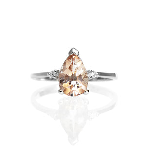 A product photo of a 9 karat white gold ring with a pear-cut peachy pink morganite centre stone sitting on a white background. The golden band is simple and smooth, meeting elegantly on either side of the morganite centre stone, hugged by a single diamond on either side.