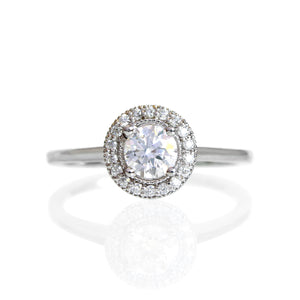 A product photo of a classic moissanite halo engagement ring in solid 9k white gold on a white background. The moissanite halo is framed by a thick band of solid 9k white gold, and the rounded band is smooth and minimalistic. The sparkling clear moissanite centre stone is held in place by 4 claws and reflects light off of its many facets. The brilliance would make it a good diamond substitute.