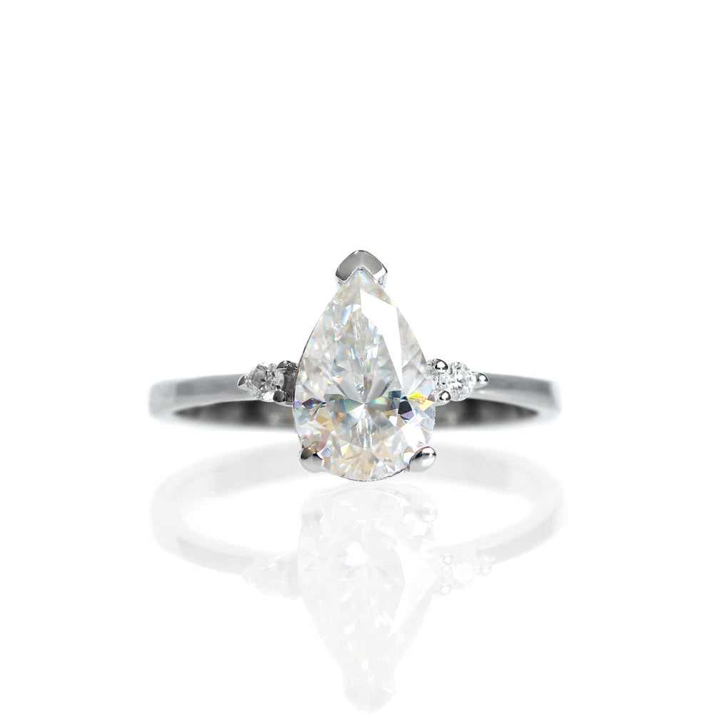 A product photo of a 9 karat white gold ring with a pear-cut clear moissanite centre stone sitting on a white background. The golden band is simple and smooth, meeting elegantly on either side of the moissanite centre stone, hugged by a single white moissanite on either side.