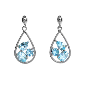 A product photo of a pair of aquamarine stud drop earrings in 9k white gold over a white background. The earrings are composed of a delicate frame of white gold in the shape of a tear, with a pear-shaped, round-shaped and oval-shaped pale blue aquamarine jewels delicately balanced atop of one another within - held in place by dainty white gold claws.