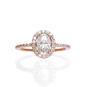 A product photo of a moissanite ring in 9k rose gold on a clear white background. A large brilliant white oval-cut jewel in the centre reflects multi-coloured light, and is framed by a ring of white moissanites. A simple round band continues the moissanite detailing for half of its length, smoothing out into a simple rose gold back.