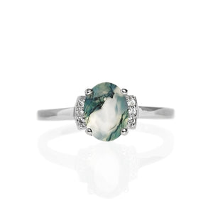 A product photo of a silver moss agate and moissanite trio ring sitting on a white background. The oval, naturally-included moss agate gemstone stands in stark contrast to the little clusters of three classic white diamond stones on either side.