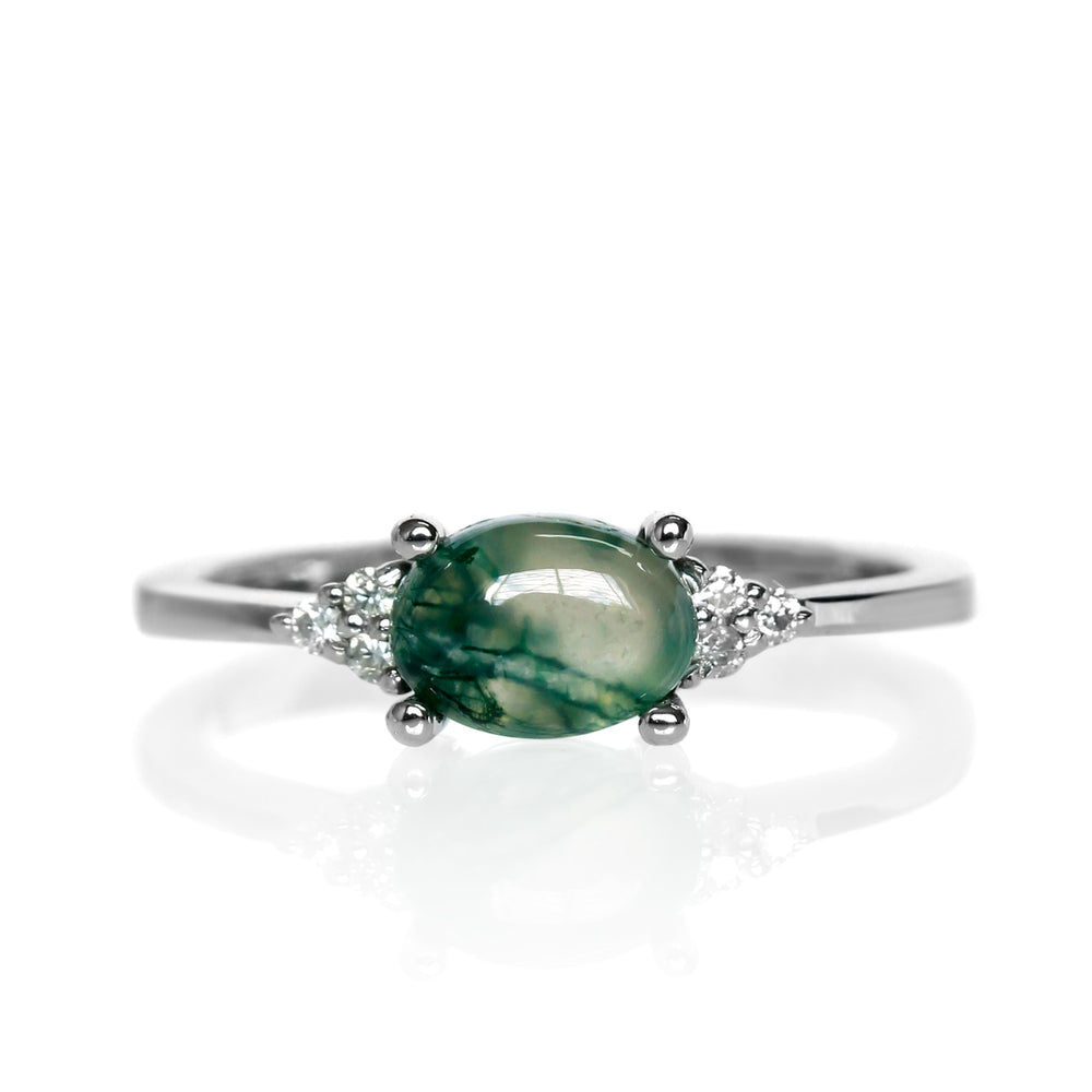 A product photo of a silver moss agate and moissanite ring sitting against a white background. The silver band is plain and smooth, and the horizontally-oriented cabochon moss agate centre stone is framed by a delicate trio of moissanite on either side.