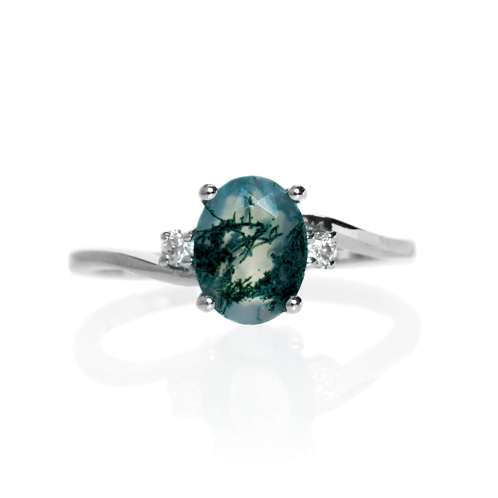 A product photo of a silver moss agate and moissanite trio ring sitting on a white background. The oval, naturally-included moss agate gemstone stands in stark contrast to the singular white moissanite stones on either side.