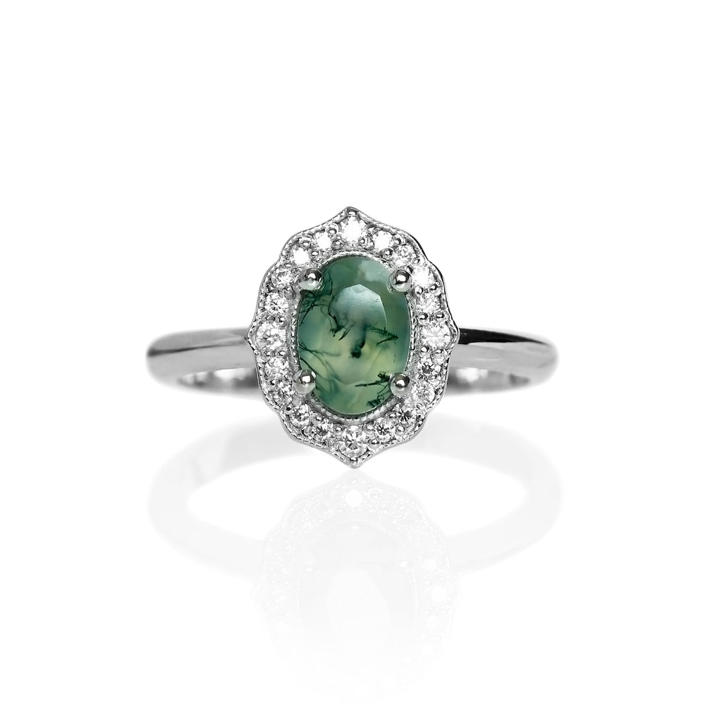 A product photo of a vintage-styled moss agate and moissanite ring in silver on a white background. The oval-shaped moss agate is held in place by 4 silver claws, and is surrounded by an ornate sterling silver frame embedded with moissanite, reminiscent of vintage jewellery styles of the past. The green gemstone colour would be a good emerald substitute.
