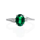 A product photo of a white gold lab emerald and lab diamond trio ring sitting on a white background. he oval, eyeclear May birthstone is a vibrant electric green, and stands in stark contrast to the singular white diamond stones on either side. The smooth 9 karat white gold band is asymmetrical, one side curving under the side diamond detail, the other curving above.