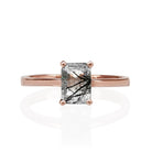 A product photo of a tourmalated quartz solitaire ring in solid 9 karat rose gold sitting on a white background. The band is simple and smooth. On the face of the ring is a vertically-oriented 7x5mm rectangular gemstone, held in place by 4 golden claws.