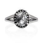 A product photo of a tourmalated quartz ring with a lavish black moissanite halo in solid sterling silver on a white background. The smooth band splits in two halfway along its length on either side, meeting the halo and tourmalated quartz centre stone in the middle. The impressively large quartz is held in place by 4 claws and is heavily included, with black striations and and strikes snaking throughout the black and white jewel.