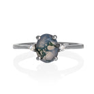 A product photo of a silver moss agate and moissanite ring sitting against a white background. The solid sterling silver band is plain and smooth, and the central oval-cut moss agate stone is framed by a single white moissanite jewel on either side.