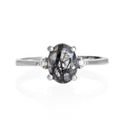 A product photo of a silver black and white tourmalated quartz and moissanite ring sitting against a white background. The solid sterling silver band is plain and smooth, and the central oval-cut monochromatic quartz stone is framed by a single white moissanite jewel on either side.