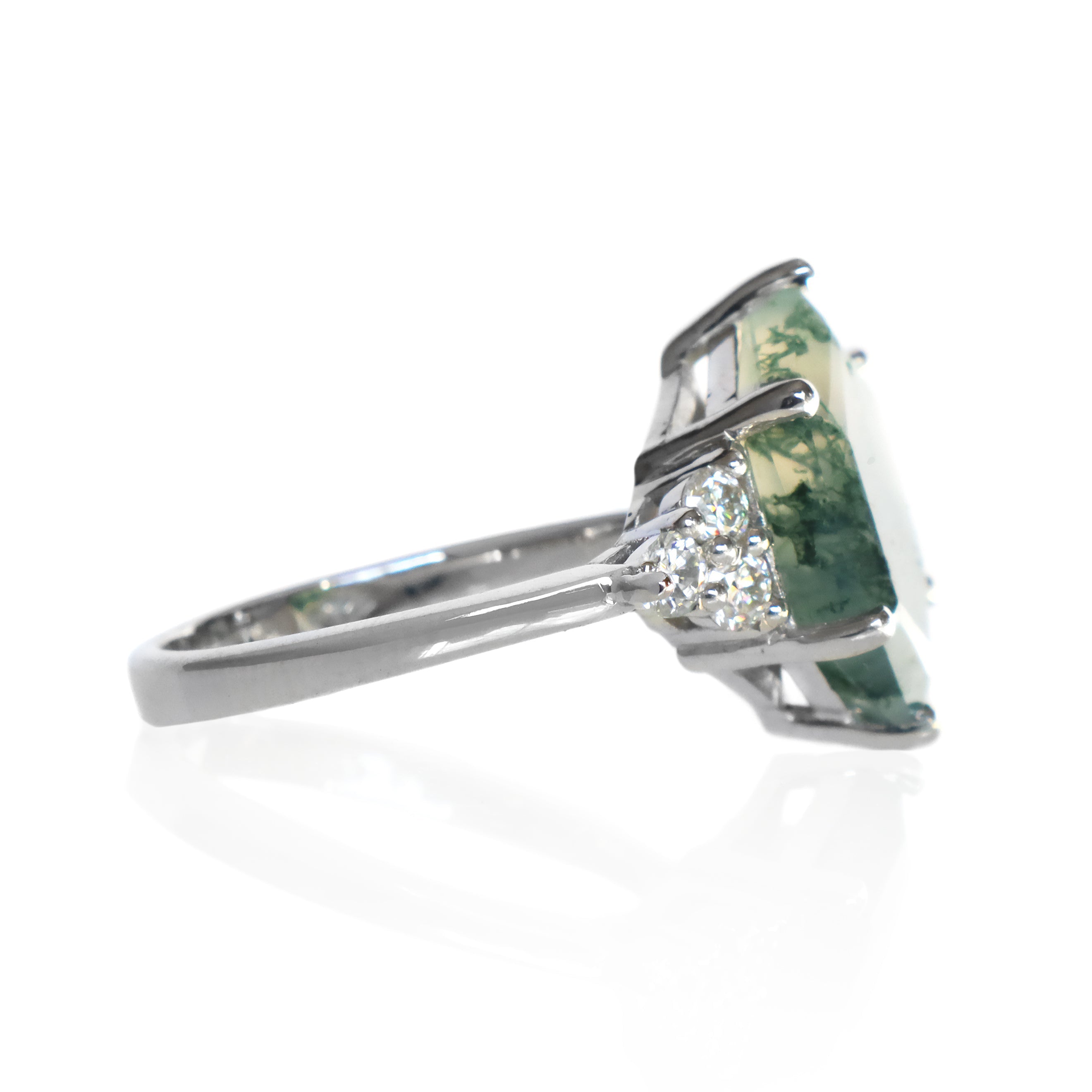 A product photo of a statement moissanite and moss agate engagement ring in solid 925 sterling silver sitting on a white background. The band is smooth and rounded. The impressively large 14x10mm Hexagonal Moss Agate gemstone is embraced on either side by a delicate trio of bright white moissanite gems.