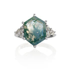 A product photo of a statement moissanite and moss agate engagement ring in solid 925 sterling silver sitting on a white background. The band is smooth and rounded. The impressively large 14x10mm Hexagonal Moss Agate gemstone is embraced on either side by a delicate trio of bright white moissanite gems.