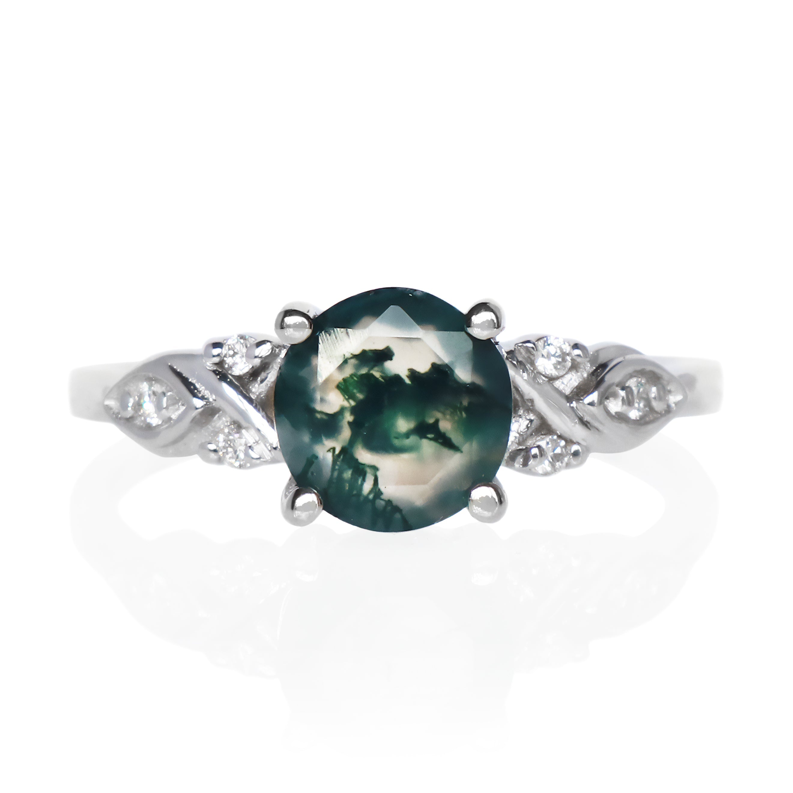 A product photo of a white gold moss agate and diamond ring sitting on a white background. The round-cut, naturally-included dark moss agate gemstone stands in stark contrast to the dazzling white diamond stones embedded in the ornate gold detailing of the ring on either side.