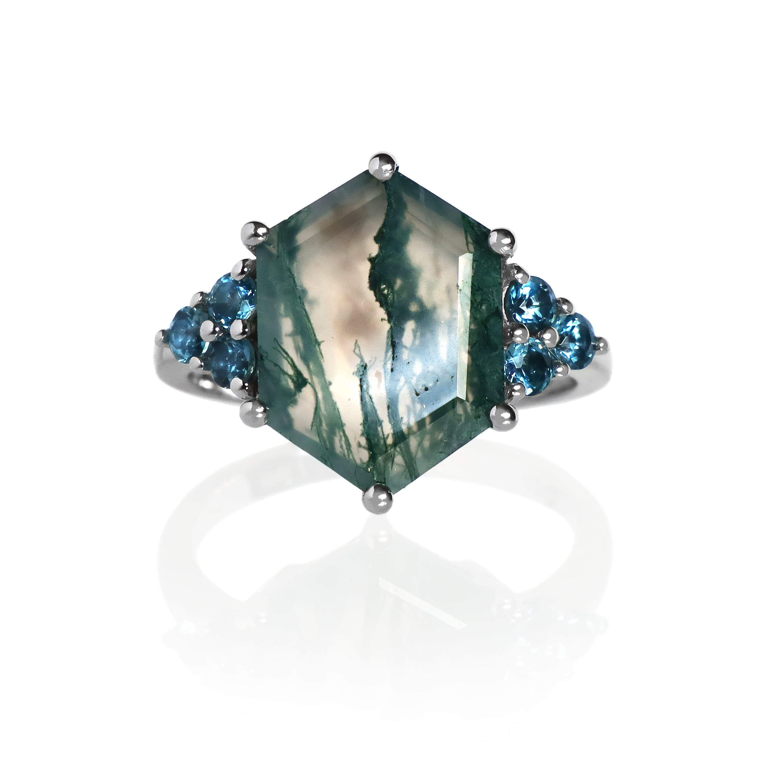 A product photo of a statement London Blue Topaz and moss agate engagement ring in solid gold sitting on a white background. The band is smooth and rounded. The impressively large 14x10mm Hexagonal Moss Agate gemstone is embraced on either side by a delicate trio of deep blue 2.5mm round London Blue Topaz gems.