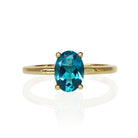 A product photo of a 9k yellow gold London blue topaz ring sitting on a white background. The ring features a remarkable 8x6mm oval-cut London blue topaz gemstone as its centerpiece, held by four polished yellow gold prongs. The London blue topaz displays a deep, rich teal-blue hue. The ring's band is a smooth, polished, and slender 9k yellow gold, offering a classic and elegant setting for the striking gemstone.
