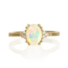 A product photo of an 8x6mm oval rainbow opal and diamond ring in 9k yellow gold, presented on a white background. The ring showcases a luminous oval-cut rainbow opal held by decorative yellow gold dual-prongs. The opal displays a vivid spectrum of color, including bright greens, blues, pinks, and oranges that shift with the light. Accentuating the central opal are clusters of three small, round brilliant-cut diamonds on each side, creating a delicate sparkle.
