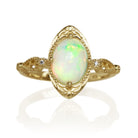 A product photo of a 9x7mm oval rainbow opal cabochon and diamond ring in 9k yellow gold, displayed on a white background. The ring features an oval-cut rainbow opal cabochon set within an elaborate, antique-inspired yellow gold frame. On each side of the central setting, the intricate 9k yellow gold band features textured openwork scroll designs, each accented with a small, round brilliant-cut diamond.