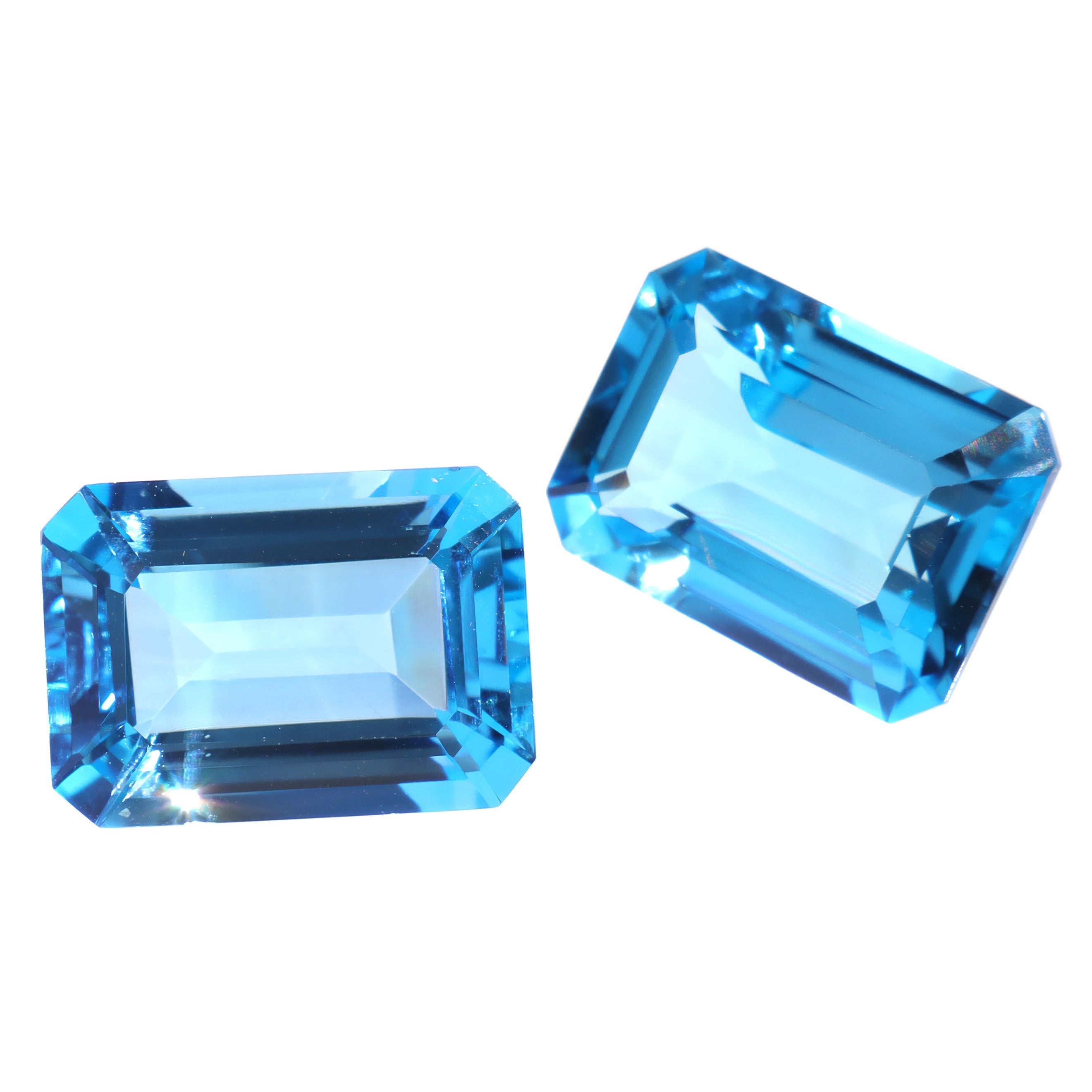 A pair of 14x10mm octagonal/emerald-cut Swiss blue topaz gemstones on a white background. The stones are perfectly-matched in both size and vibrant blue hue, and are ideal for use in a bespoke pair of birthstone earrings, or as a gift to two people with a November birthday.