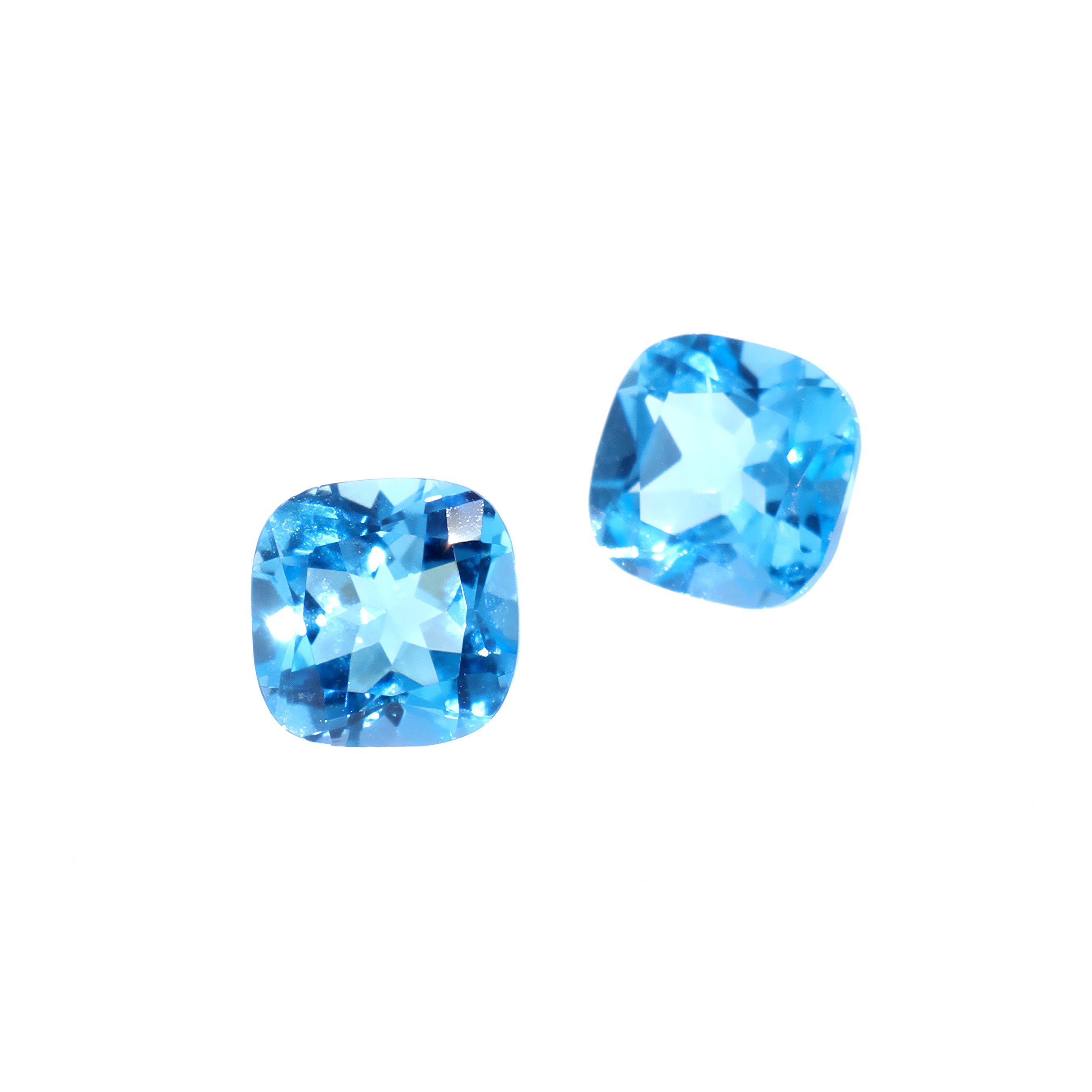 A pair of loose cushion-cut Swiss blue topaz gemstones sitting on a white background. The stones are elaborately faceted, showing off their vibrant blue colouring and fire throughout. The stones would be perfect for use in a bespoke custom-designed ring and pendant set, or as a pair of November birthstone earrings.
