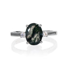 A product photo of an elegant sterling silver ring with a stunning trio of moss agate and moissanites sitting against a white background. The largest of the stones sits in the centre, an oval moss agate - hugged by two smaller circular white moissanites on either side.