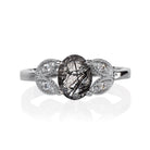 A product photo of an oval tourmalated quartz and moissanite ring with floral silver detailing set in solid 925 sterling silver metal. The bold 8x6mm tourmalated quartz gemstone is held in place by two petal shapes in filigree silver detailing on either side, each "petal" adorned with a single moissanite in the centre, to a total of 4.