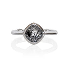 A product photo of a silver ring with a bezel-set tourmalated quartz centre stone sitting on a white background. The silver band is simple and smooth, connecting on either side of a diagonally-oriented squared-cushion tourmalated quartz stone surrounded by a solid frame of silver.