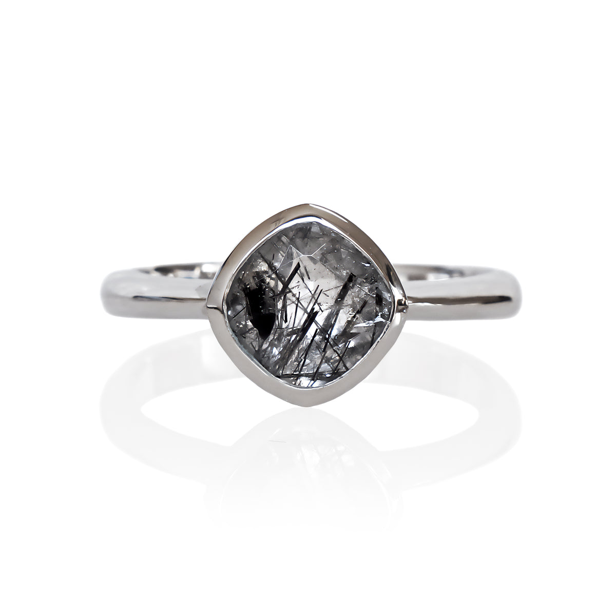 A product photo of a silver ring with a bezel-set tourmalated quartz centre stone sitting on a white background. The silver band is simple and smooth, connecting on either side of a diagonally-oriented squared-cushion tourmalated quartz stone surrounded by a solid frame of silver.