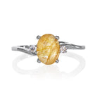 A product photo of a silver golden rutilated quartz and moissanite trio ring sitting on a white background. The oval, naturally-included golden rutilated quartz gemstone stands in stark contrast to the singular white moissanite stones on either side.