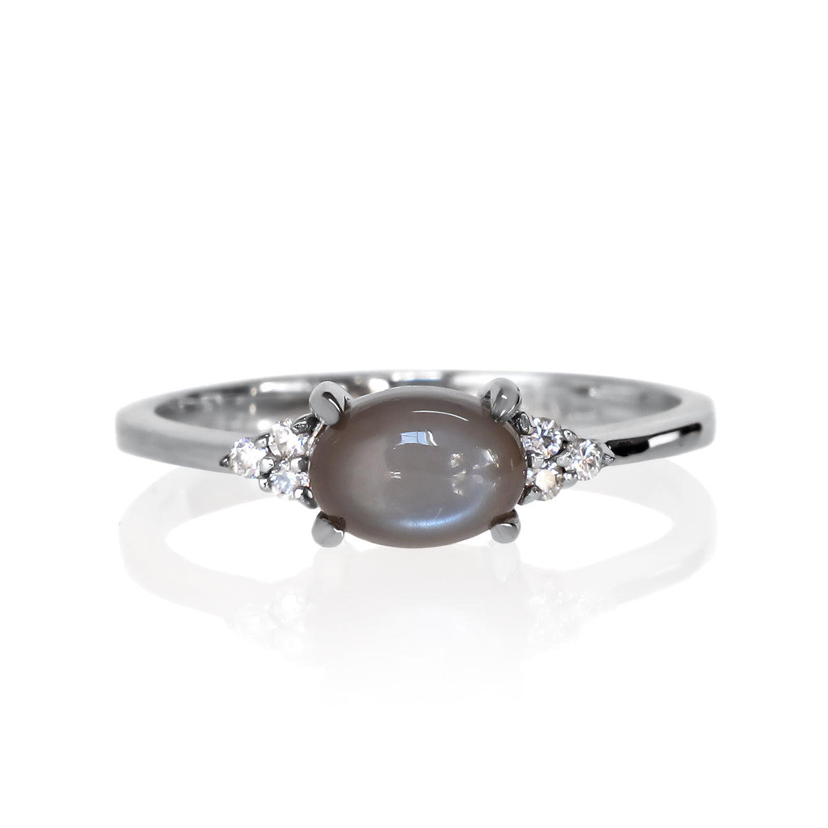 A product photo of a silver grey moonstone and moissanite ring sitting against a white background. The silver band is plain and smooth, and the horizontally-oriented cabochon moonstone centre stone is framed by a delicate trio of moissanite on either side.