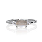 A product photo of a silver moonstone and moissanite ring sitting against a white background. The silver band is plain and smooth, and the horizontally-oriented cabochon moonstone centre stone is framed by a delicate trio of moissanite on either side.