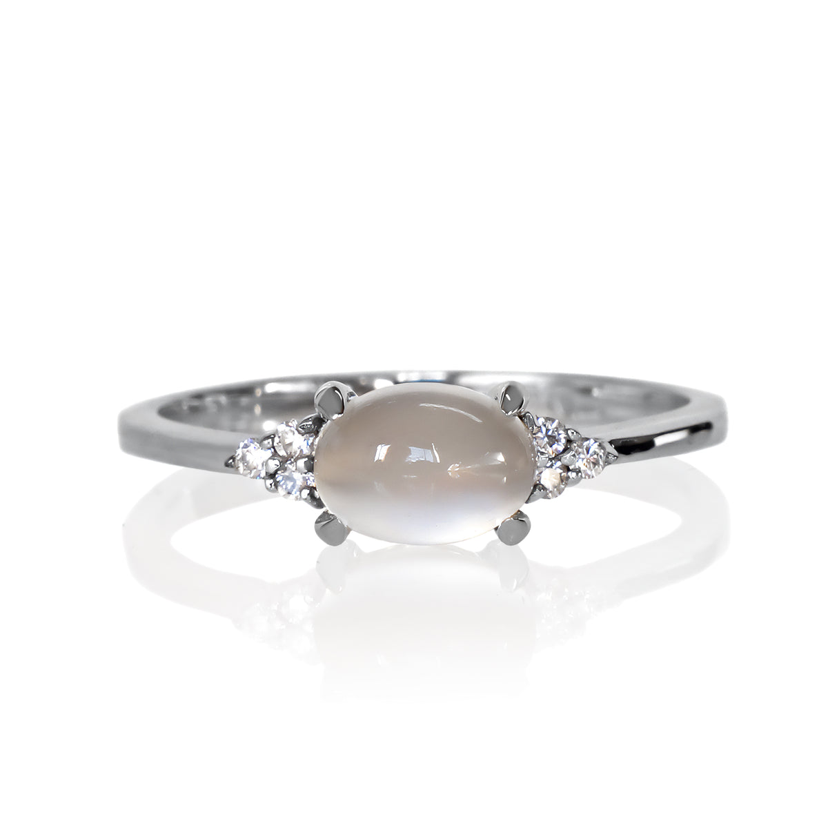 A product photo of a silver moonstone and moissanite ring sitting against a white background. The silver band is plain and smooth, and the horizontally-oriented cabochon moonstone centre stone is framed by a delicate trio of moissanite on either side.