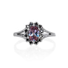 A product photo of a lavish floral moissanite halo lab alexandrite ring in solid silver on a white background. The smooth band splits in two halfway along its length on either side, meeting the floral halo and lab alexandrite in the centre. The lab alexandrite centre stone is held in place by 4 claws and is lightly included, with natural black strikes snaking throughout the monochromatic jewel.